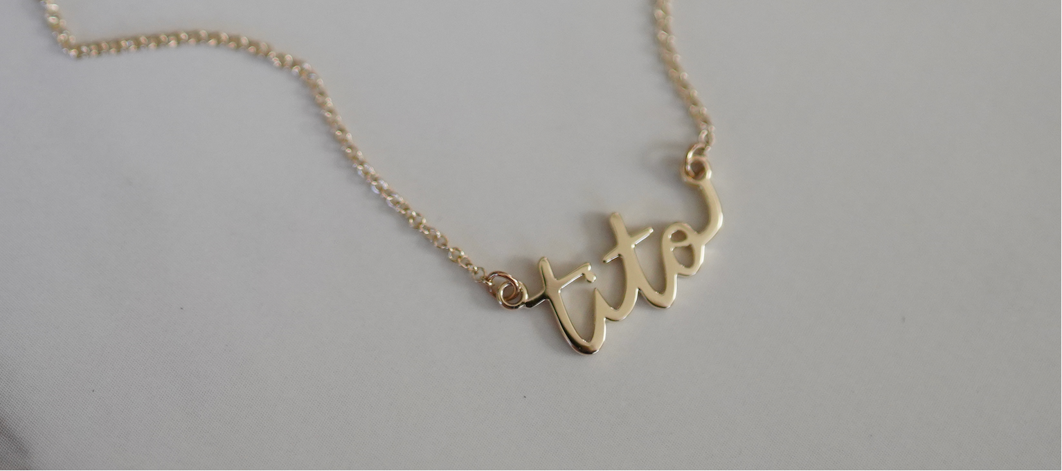 A gold script name necklace spelling 'tito' on a delicate chain, displayed on a neutral light gray background.